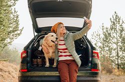 A blond woman sits on the open tailgate of her car with her golden retriever. The woman is taking a selfie of herself and her dog.