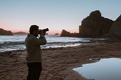 A man is standing on a beach at sunset. He is holding a camera and taking a photograph of the scenery.