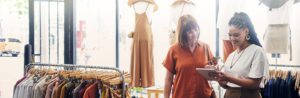 Two women are in a retail clothing store surrounded by racks of clothing on hangers.
