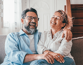 A man and woman sit on a sofa laughing. He has his arm around her shoulder.