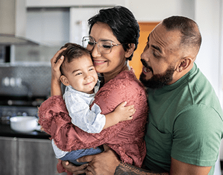 Image of a woman holding a small toddler closely. Her husband is peeking over her shoulder smiling at the child.